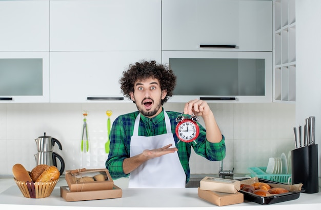 Vista frontal del hombre sorprendido de pie detrás de la mesa varios pasteles y apuntando el reloj en la cocina blanca