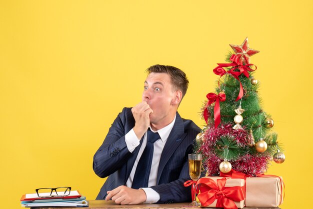 Vista frontal del hombre sorprendido sentado en la mesa cerca del árbol de Navidad y presenta en la pared amarilla espacio libre