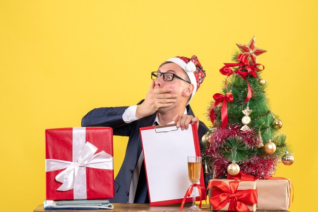 Vista frontal del hombre sorprendido sentado en la mesa cerca del árbol de Navidad y presenta en la pared amarilla espacio libre