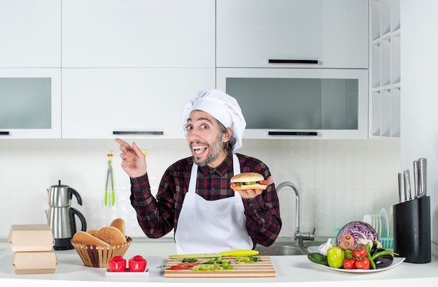Vista frontal del hombre sorprendido sosteniendo una hamburguesa de pie detrás de la mesa de la cocina