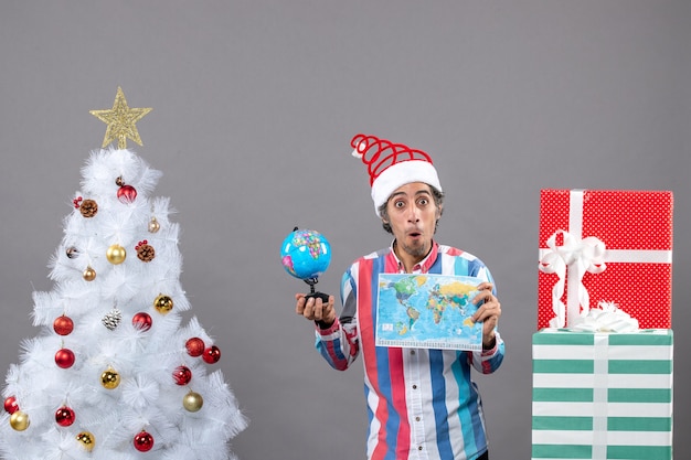 Vista frontal hombre sorprendido sosteniendo mapa del mundo y globo cerca del árbol de navidad y cajas de regalo