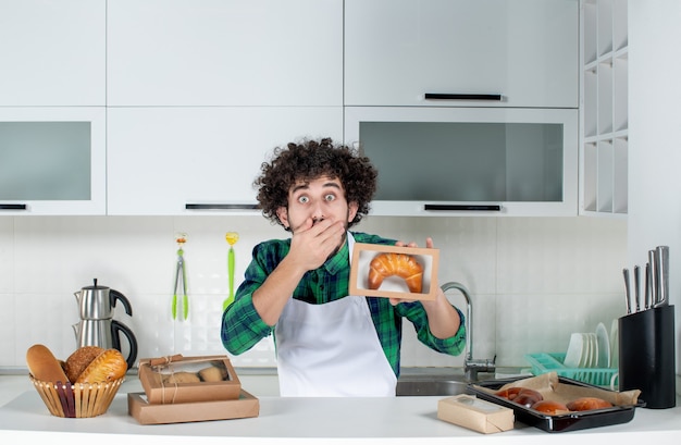 Vista frontal del hombre sorprendido sosteniendo pasteles recién horneados en una pequeña caja en la cocina blanca