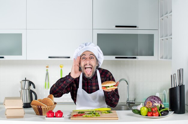 Vista frontal del hombre sosteniendo una hamburguesa de pie detrás de la mesa de la cocina