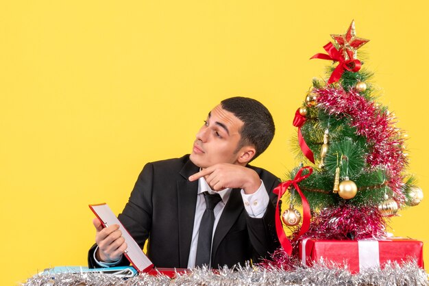 Vista frontal del hombre en traje sentado en la mesa mirando a la derecha sosteniendo el documento árbol de Navidad y regalos