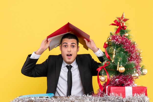 Vista frontal del hombre en traje sentado en la mesa poniendo documentos sobre su cabeza árbol de Navidad y regalos