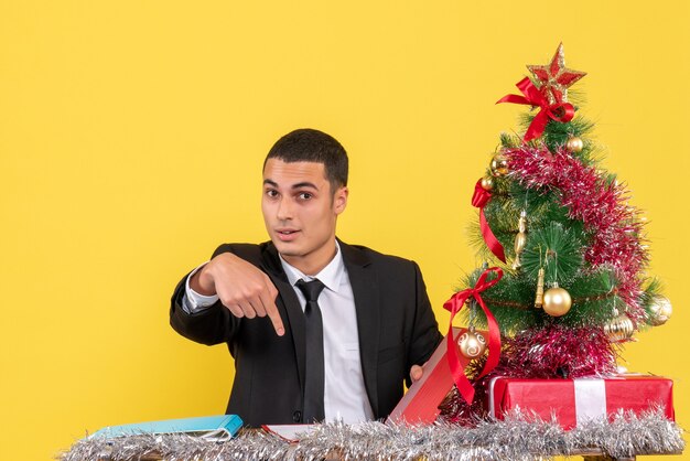 Vista frontal del hombre en traje sentado en la mesa sosteniendo el documento apuntando con el dedo hacia abajo árbol de Navidad y regalos