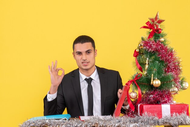 Vista frontal del hombre en traje sentado en la mesa sosteniendo el documento haciendo okey firmar árbol de Navidad y regalos