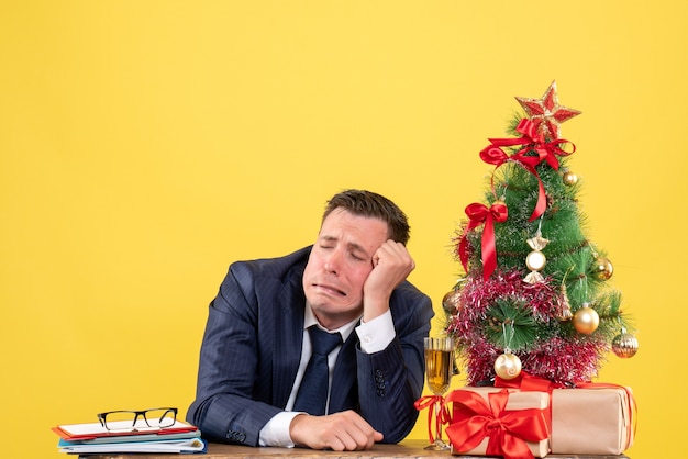 Vista frontal del hombre triste sentado en la mesa cerca del árbol de Navidad y presenta en amarillo