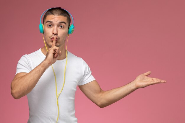 Vista frontal de los hombres jóvenes en camiseta blanca escuchando música que muestra el signo de silencio sobre fondo rosa claro