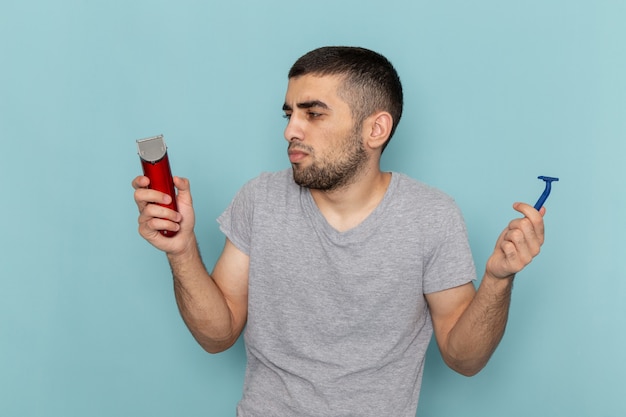 Vista frontal de los hombres jóvenes en camiseta gris sosteniendo una maquinilla de afeitar eléctrica roja y una maquinilla de afeitar simple en la barba azul hielo espuma afeitadora de pelo