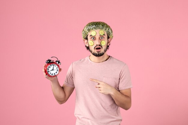 Vista frontal de los hombres jóvenes con máscara en la cara sosteniendo el reloj sobre fondo de color rosa