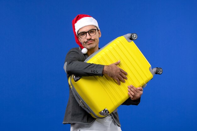 Vista frontal de los hombres jóvenes que llevan una pesada bolsa amarilla en las vacaciones de avión de vuelo de pared azul