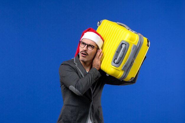 Vista frontal de los hombres jóvenes que llevan una pesada bolsa amarilla en las vacaciones de avión de vuelo de pared azul