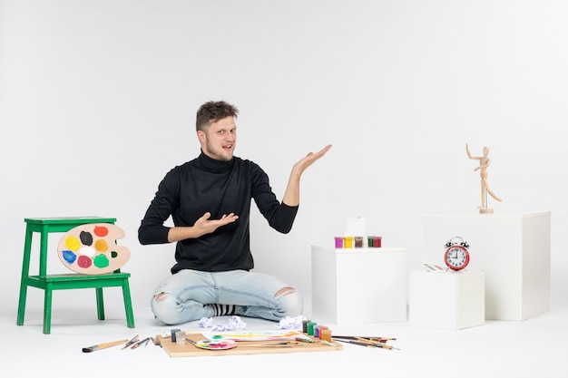 Vista frontal de los hombres jóvenes sentados alrededor de pinturas y borlas para dibujar en el arte de la pared blanca dibujar pintura color artista