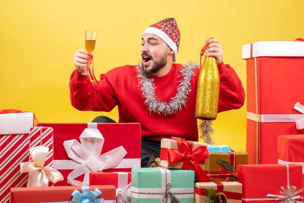 Vista frontal de los hombres jóvenes sentados alrededor de regalos celebrando con champán sobre fondo amarillo