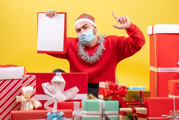 Vista frontal de los hombres jóvenes sentados alrededor de los regalos de Navidad con una nota sobre fondo amarillo