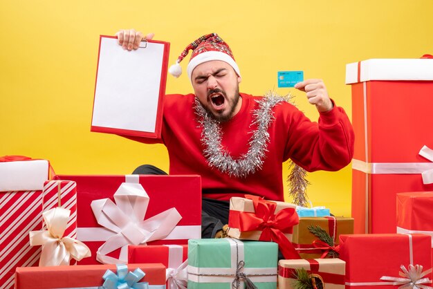 Vista frontal de los hombres jóvenes sentados alrededor de los regalos de Navidad sobre fondo amarillo