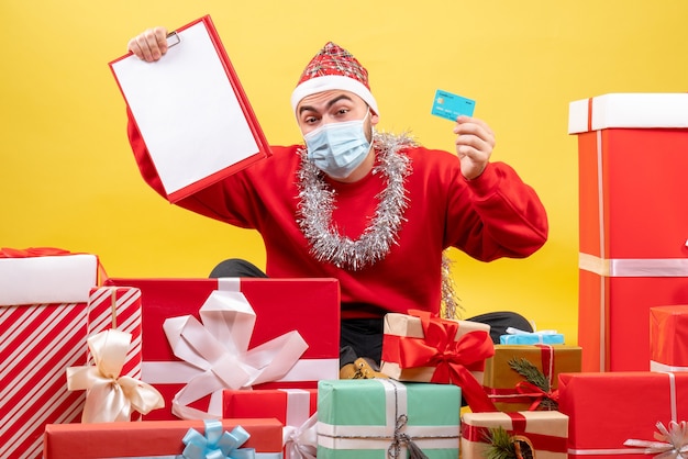 Vista frontal de los hombres jóvenes sentados alrededor de regalos con nota y tarjeta bancaria sobre fondo amarillo