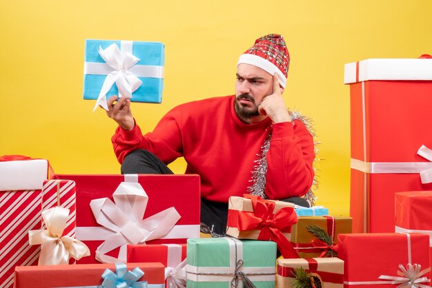 Vista frontal de los hombres jóvenes sentados alrededor de regalos y sosteniendo uno sobre fondo amarillo