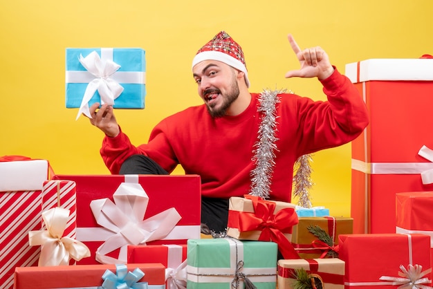 Vista frontal de los hombres jóvenes sentados alrededor de regalos y sosteniendo uno sobre fondo amarillo