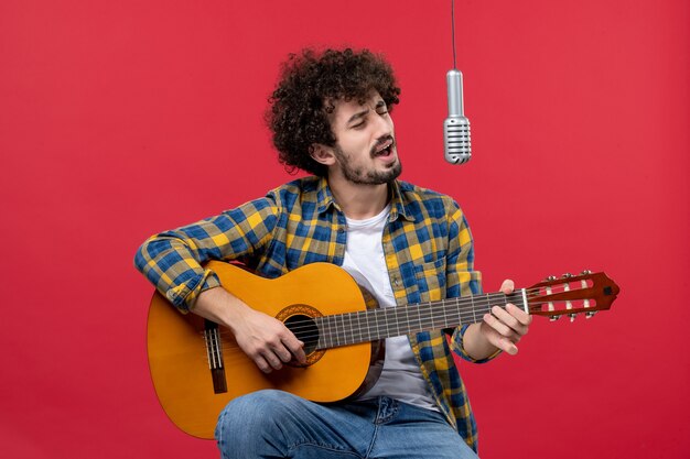 Vista frontal de los hombres jóvenes sentados con la guitarra y cantando en la pared roja color aplauso músico tocar música de concierto en vivo