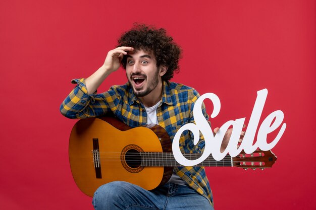 Vista frontal de los hombres jóvenes sentados con la guitarra en la pared roja color aplauso músico tocar la banda concierto venta en vivo
