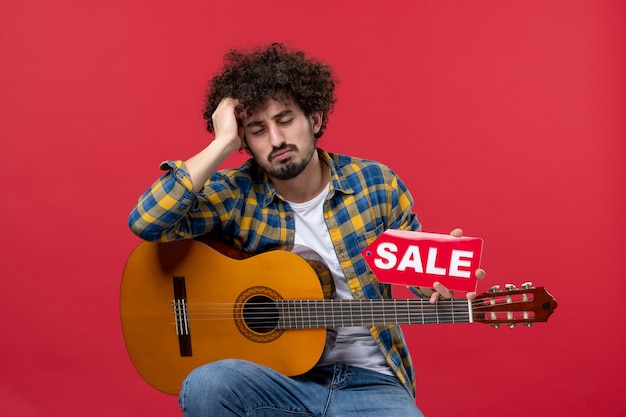 Vista frontal de los hombres jóvenes sentados con la guitarra en la pared roja color aplauso músico tocar música de banda venta en vivo