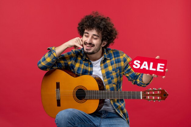 Vista frontal de los hombres jóvenes sentados con la guitarra en la pared roja color aplauso músico en vivo concierto venta play play