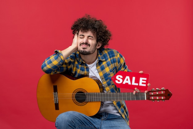 Vista frontal de los hombres jóvenes sentados con la guitarra en la pared roja concierto colores vivos aplauso músico venta reproducir música