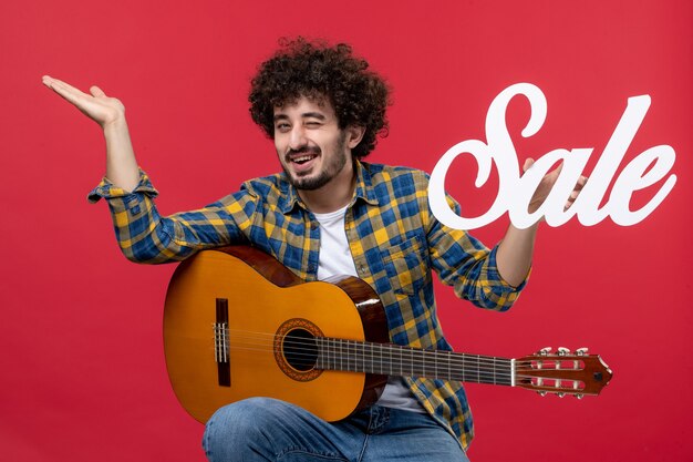 Vista frontal de los hombres jóvenes sentados con la guitarra en la pared roja concierto de música músico en vivo venta de colores