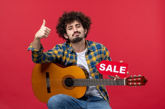 Vista frontal de los hombres jóvenes sentados con la guitarra en la pared roja concierto música en vivo aplauso de color músico venta