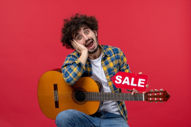 Vista frontal de los hombres jóvenes sentados con la guitarra en la pared roja concierto músico en vivo venta reproducir música aplauso de color