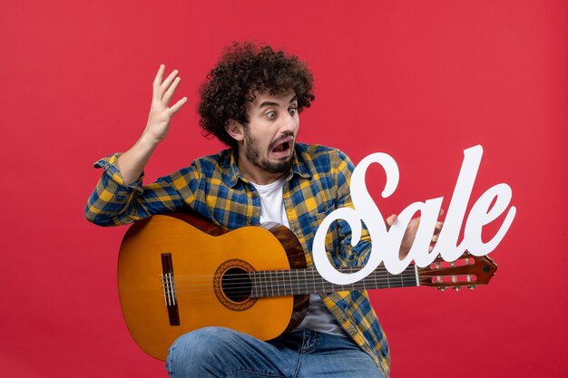 Vista frontal de los hombres jóvenes sentados con la guitarra en la pared roja concierto músico en vivo venta reproducir música color