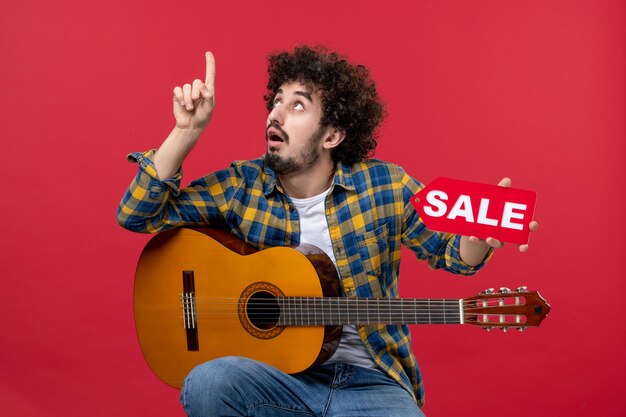 Vista frontal de los hombres jóvenes sentados con la guitarra en la pared roja música color aplauso músico en vivo venta play