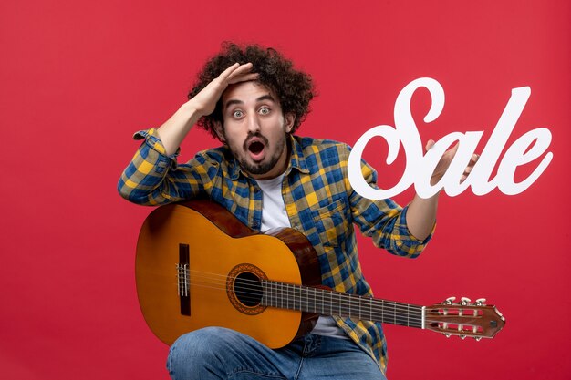 Vista frontal de los hombres jóvenes sentados con la guitarra en la pared roja música concierto aplauso músico venta de colores