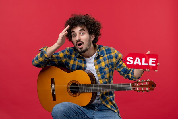 Vista frontal de los hombres jóvenes sentados con la guitarra en la pared roja reproducir música músico color concierto en vivo venta