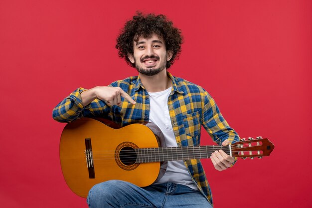 Vista frontal de los hombres jóvenes sentados con la guitarra en la pared roja, tocar el concierto en vivo de la banda, músico de color, aplausos