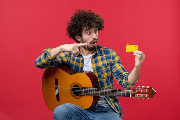 Vista frontal de los hombres jóvenes sentados con la guitarra sosteniendo una tarjeta bancaria en la pared roja, concierto de música, músico, aplauso, color vivo