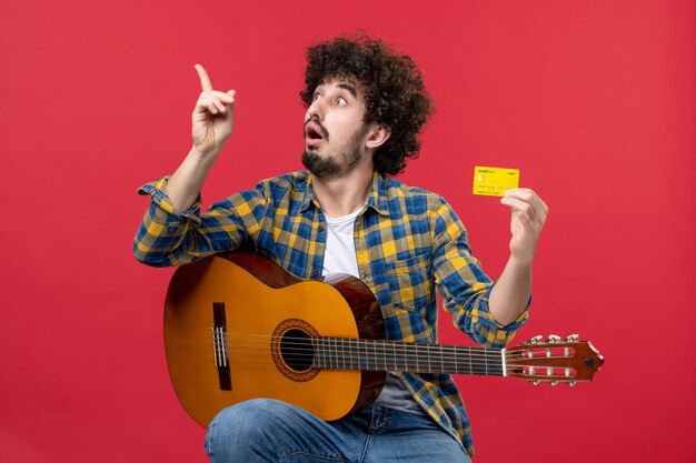 Vista frontal de los hombres jóvenes sentados con la guitarra sosteniendo una tarjeta bancaria en la pared roja, concierto de música, músico, aplauso, color vivo