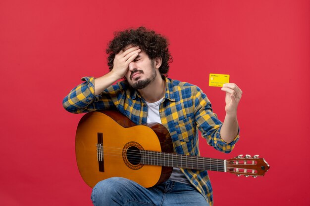 Vista frontal de los hombres jóvenes sentados con la guitarra sosteniendo una tarjeta bancaria en la pared roja, concierto de rendimiento de color, aplausos, música en vivo