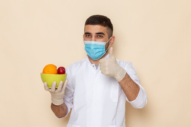 Vista frontal de los hombres jóvenes en traje blanco con máscara y guantes que sostienen el plato con frutas en beige