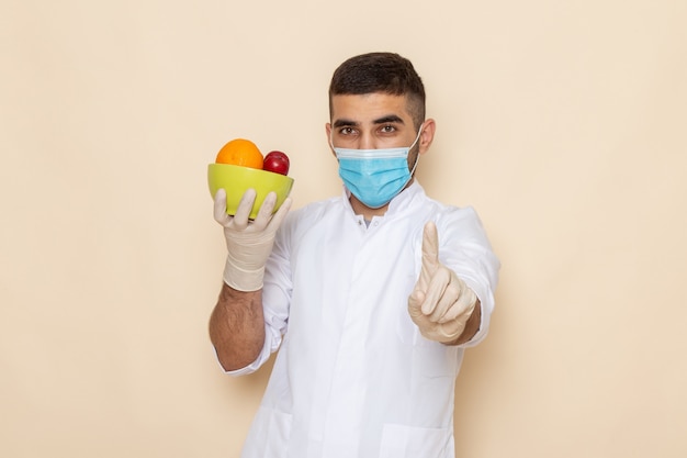 Vista frontal de los hombres jóvenes en traje blanco con máscara y guantes que sostienen el plato con frutas en beige