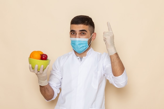 Vista frontal de los hombres jóvenes en traje blanco con máscara y guantes que sostienen el plato con frutas en beige