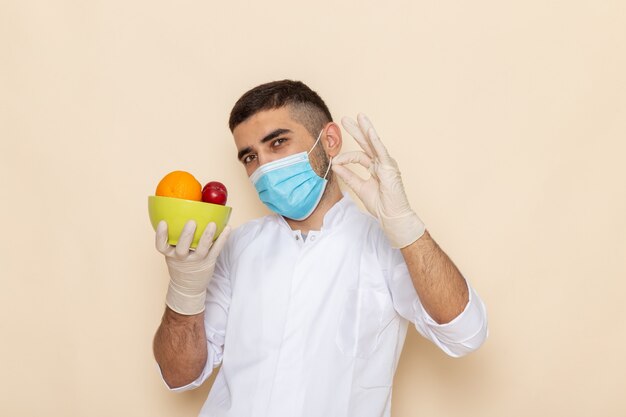 Vista frontal de los hombres jóvenes en traje blanco con máscara y guantes que sostienen el plato con frutas en beige