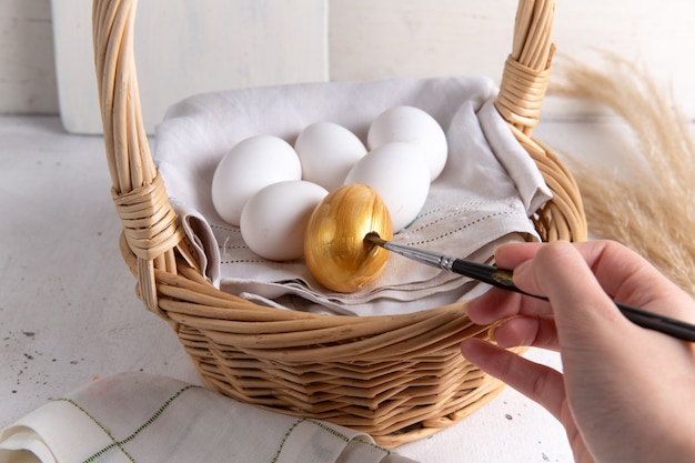 Vista frontal de huevos enteros blancos dentro de la canasta con huevo dorado se pinta sobre el escritorio blanco.