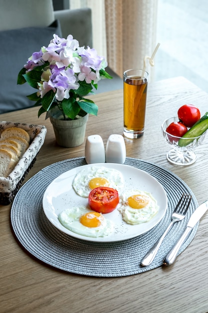 Vista frontal huevos fritos con tomate en un plato con un refresco y flores en una olla sobre la mesa