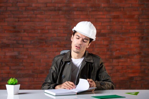 Vista frontal ingeniero masculino sentado detrás de su lugar de trabajo en el plan de documento de bloc de notas de lista de casco blanco plan de propiedad de contratista de constructor de trabajo corporativo de negocios