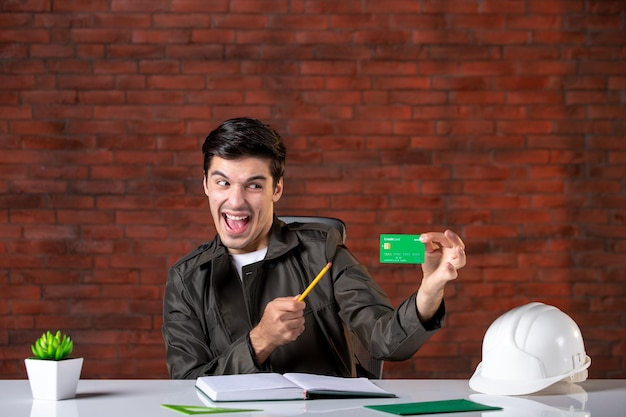 Vista frontal ingeniero masculino sentado detrás de su lugar de trabajo y sosteniendo plan de construcción de tarjeta de crédito verde agenda de propiedad comercial de contratista corporativo