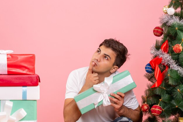 Vista frontal del joven alrededor de regalos y árbol de Navidad en la pared rosa