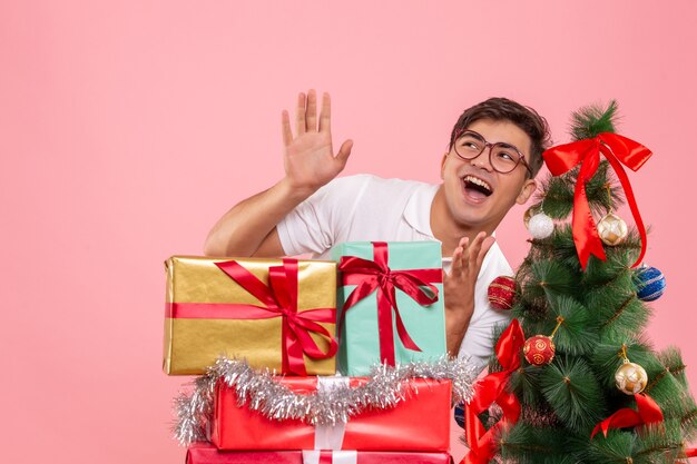 Vista frontal del joven alrededor de regalos de Navidad y árbol de vacaciones en pared rosa